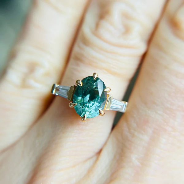 14K yellow gold set with a 2.56ct bi-colour, blue-green, sapphire centre. With 0.36tcw of lab grown, tapered baguette cut white-diamonds set on the sides. Worn on hand.