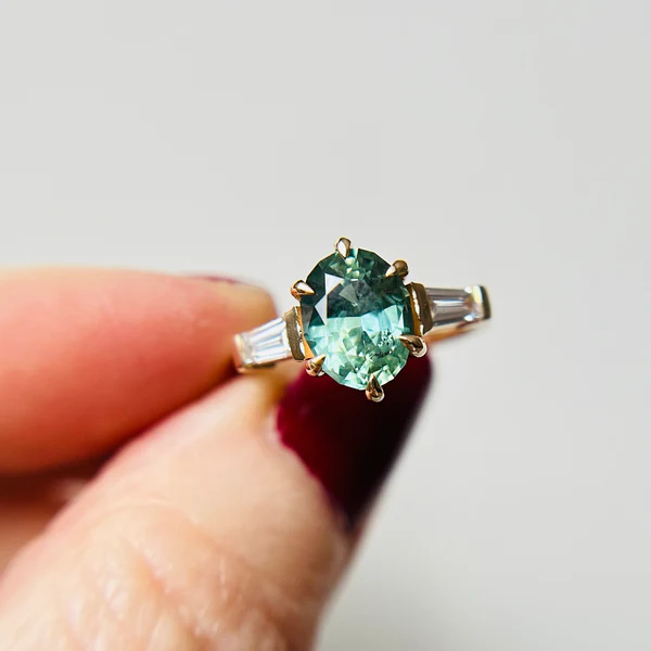 14K yellow gold set with a 2.56ct bi-colour, blue-green, sapphire centre.
With 0.36tcw of lab grown, tapered baguette cut white-diamonds set on the sides.