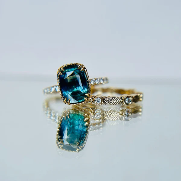 14k yellow gold ring set with sapphire cushion cut and accent white diamonds. Shown with a wedding band with diamonds and leaf engraving.