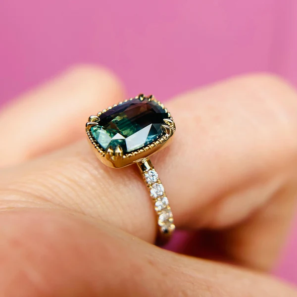 14k yellow gold ring set with sapphire cushion cut and accent white diamonds. Shown on a hand.