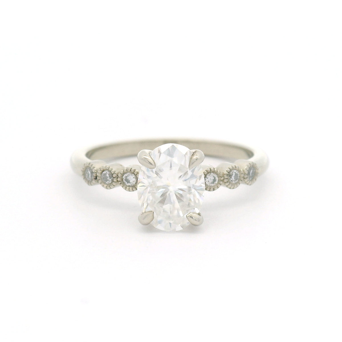 10K white gold engagement ring with a 1.50ct oval moissanite, six bezel-set round brilliant-cut moissanites and fine milgrain details.