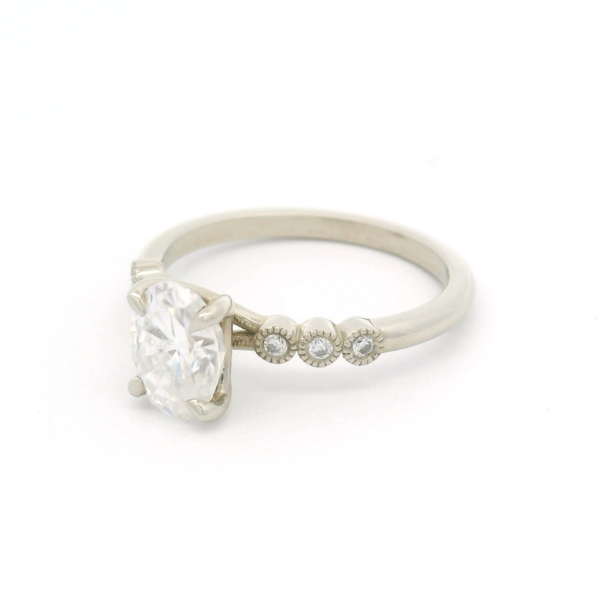 10K white gold engagement ring with a 1.50ct oval moissanite, six bezel-set round brilliant-cut moissanites and fine milgrain details.