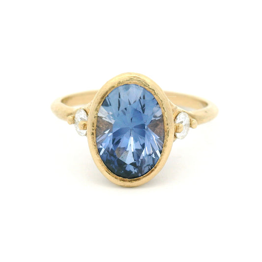 10K yellow gold organic three-stone engagement ring with a bezel-set 3ct lab-grown oval blue sapphire and two claw-set moissanite shoulder stones.