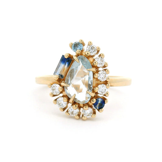 14K yellow gold asymmetrical engagement ring featuring a natural 0.65ct triangular greyish-blue sapphire with a halo of mixed-cut blue sapphires and lab-grown diamonds totalling 0.26cts.