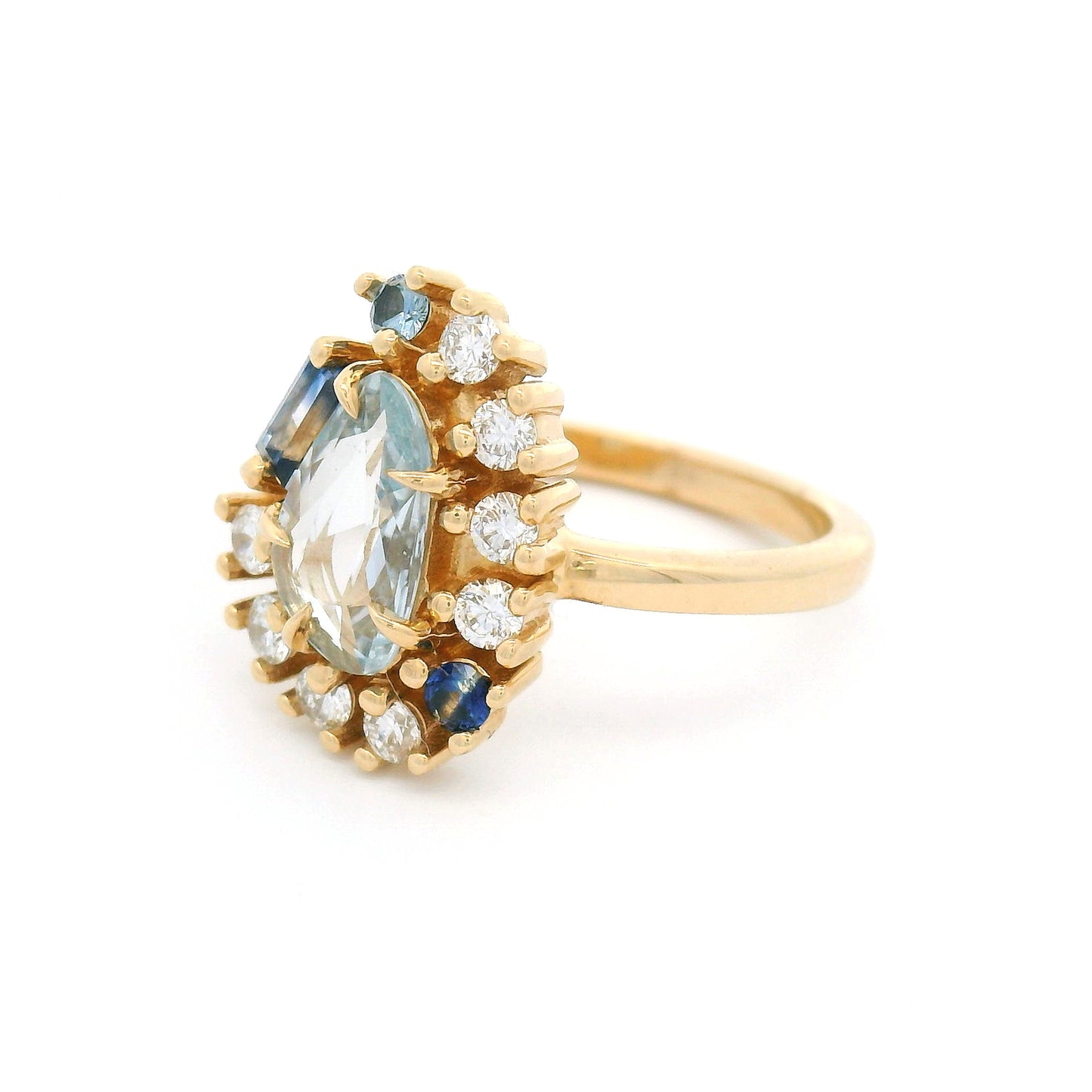 14K yellow gold asymmetrical engagement ring featuring a natural 0.65ct triangular greyish-blue sapphire with a halo of mixed-cut blue sapphires and lab-grown diamonds totalling 0.26cts.