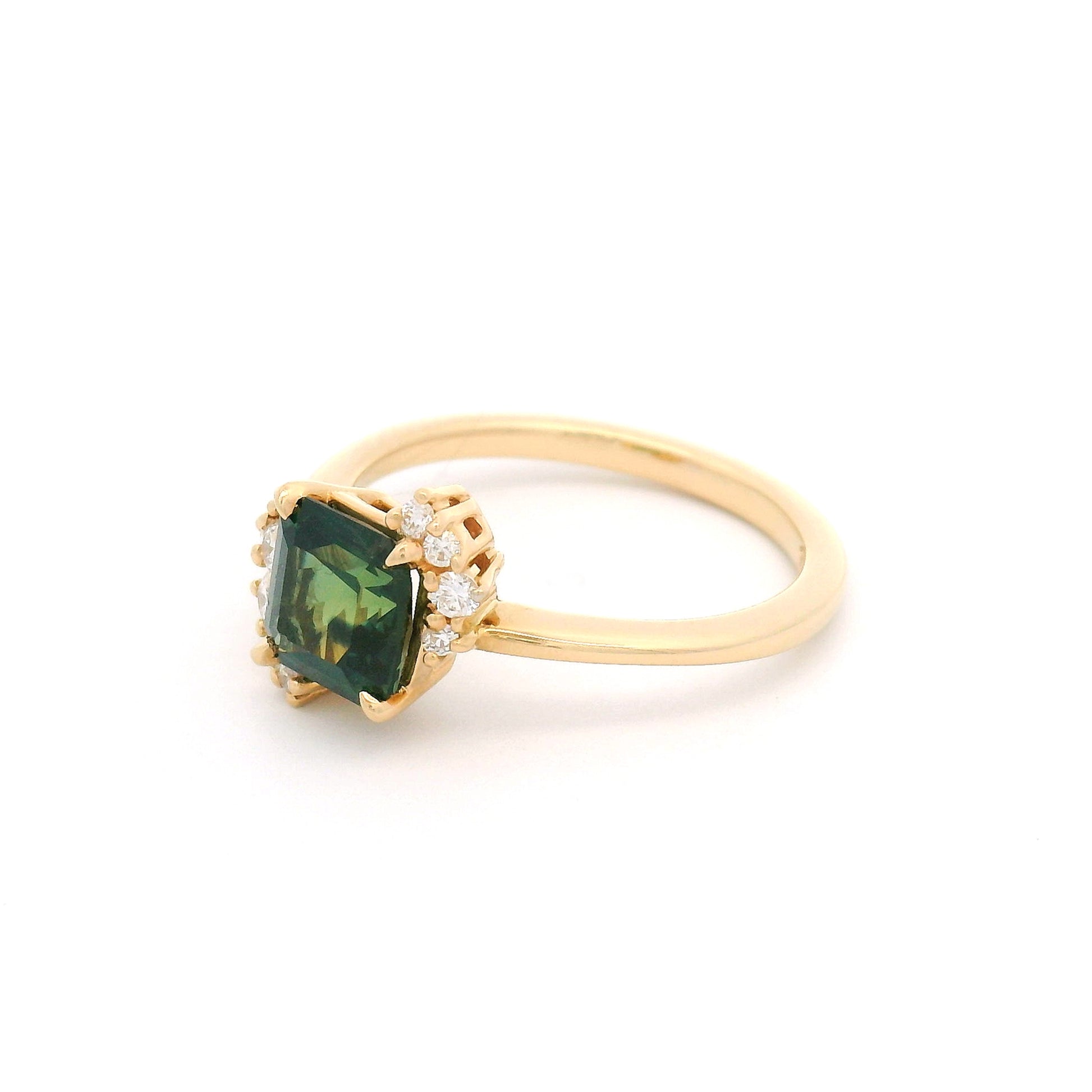 14K yellow gold asymmetrical engagement ring featuring a claw-set natural 1.41ct square radiant-cut dark green-teal sapphire with 10 claw-set natural diamond accents totalling 0.16cts on a delicate 1.7mm band.