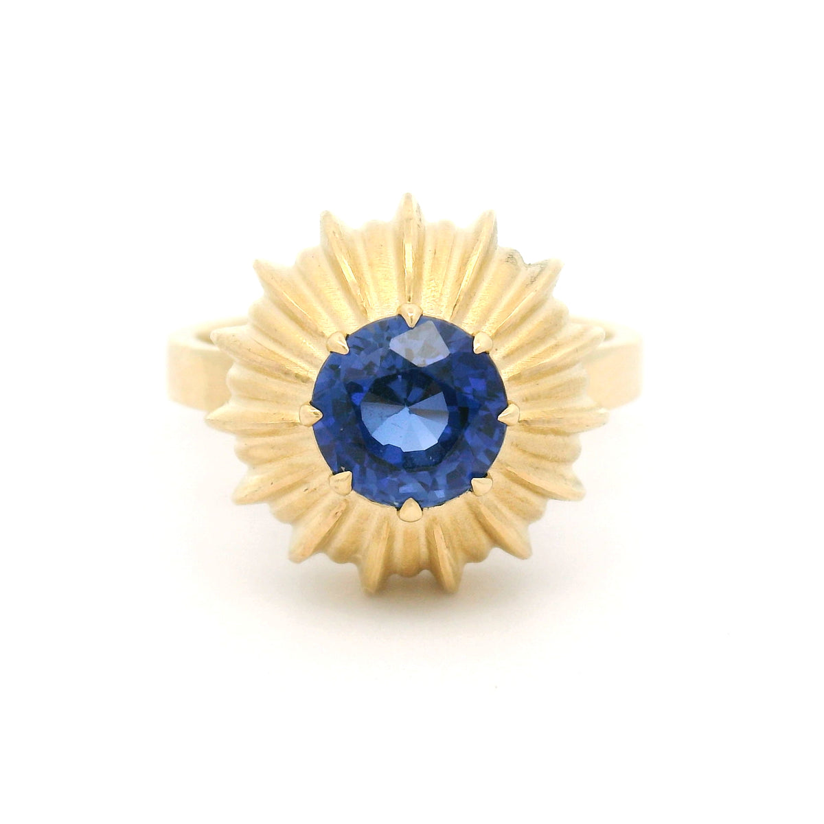 14K yellow gold claw-set solitaire engagement ring featuring a 6.6mm lab-grown round blue sapphire in a sculptural fluted sunburst setting.