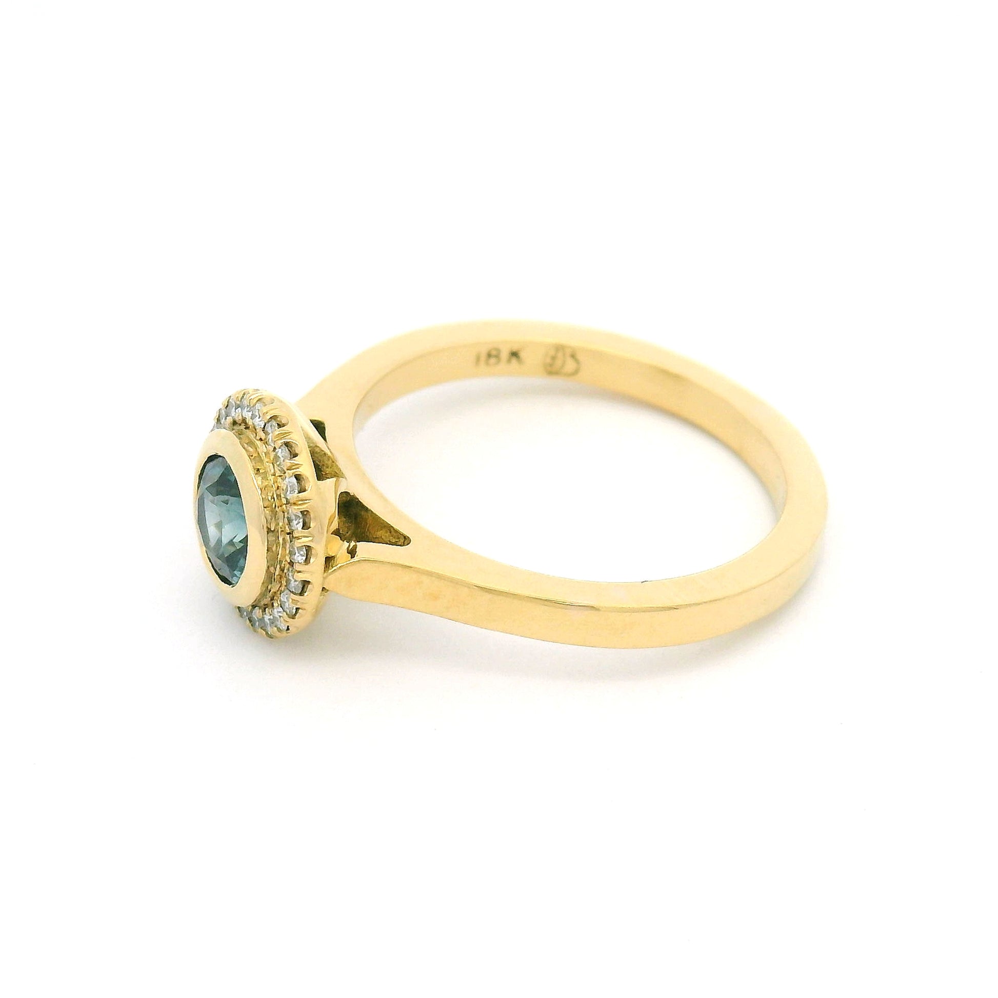 18K yellow gold engagement ring with a bezel-set natural 0.65ct round teal sapphire and bead-set halo of 0.12cts natural diamonds.